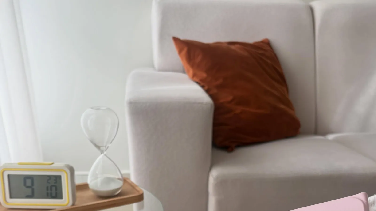 Canto do consultório com sofá claro, almofada cor de terra, uma ampulheta e um relógio sobre a mesa lateral