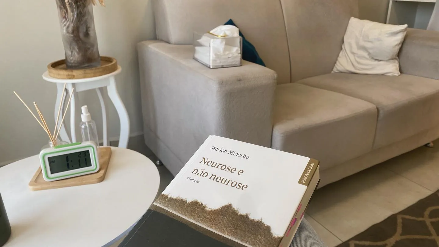 Sofá claro do consultório, com um relógio de mesa e o livro “Neurose e não neurose” em primeiro plano